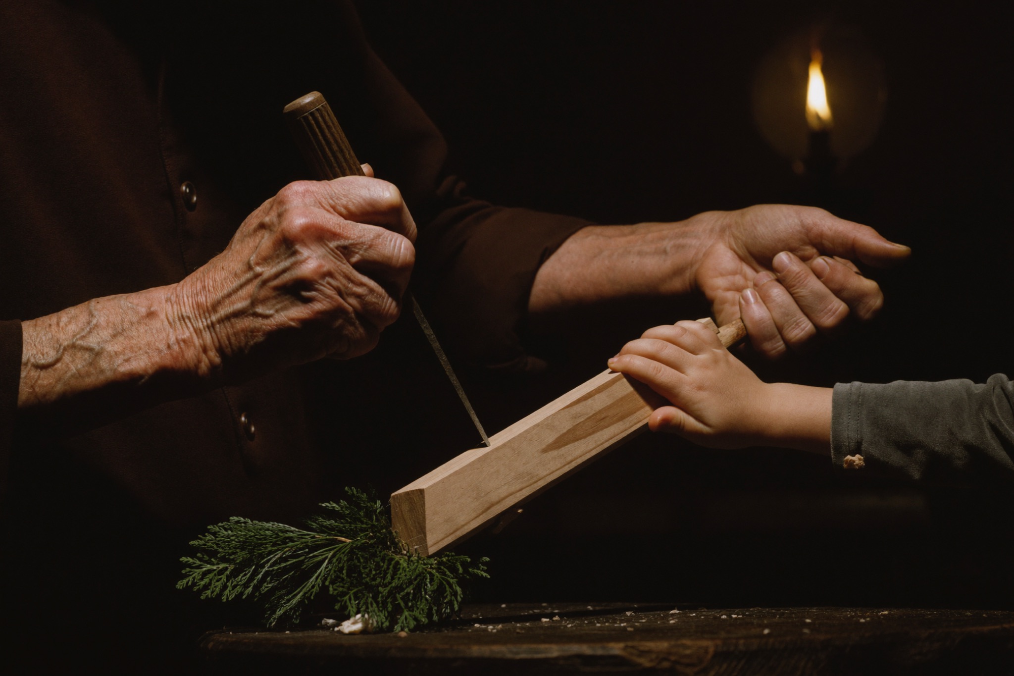 An old clockmaker's weathered hands guiding a child's hands over a linden carving, one chisel held together, by candlelight